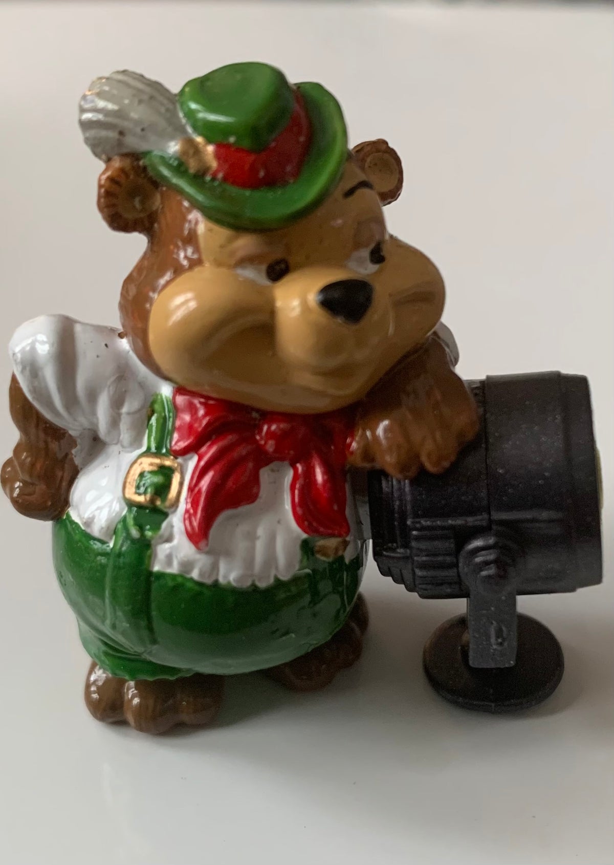 A brown bear figurine wearing a green hat with white feather red scarf and green overalls holding a black spotlight