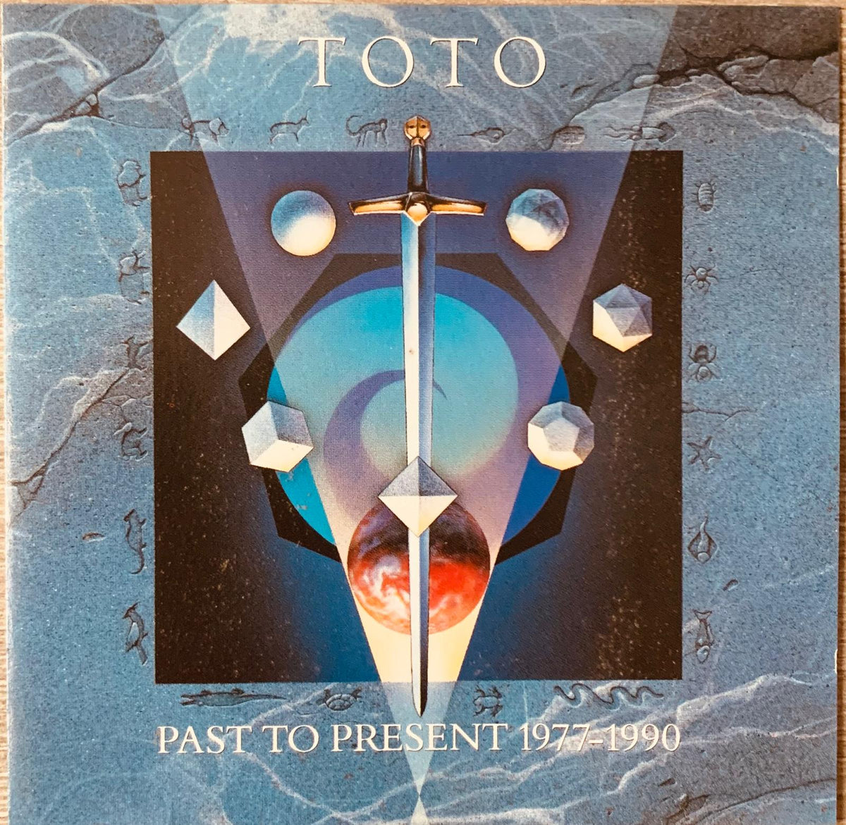 Toto original cd past to present 1977-1990 with a silver sword and ornate golden hilt on a cosmic background