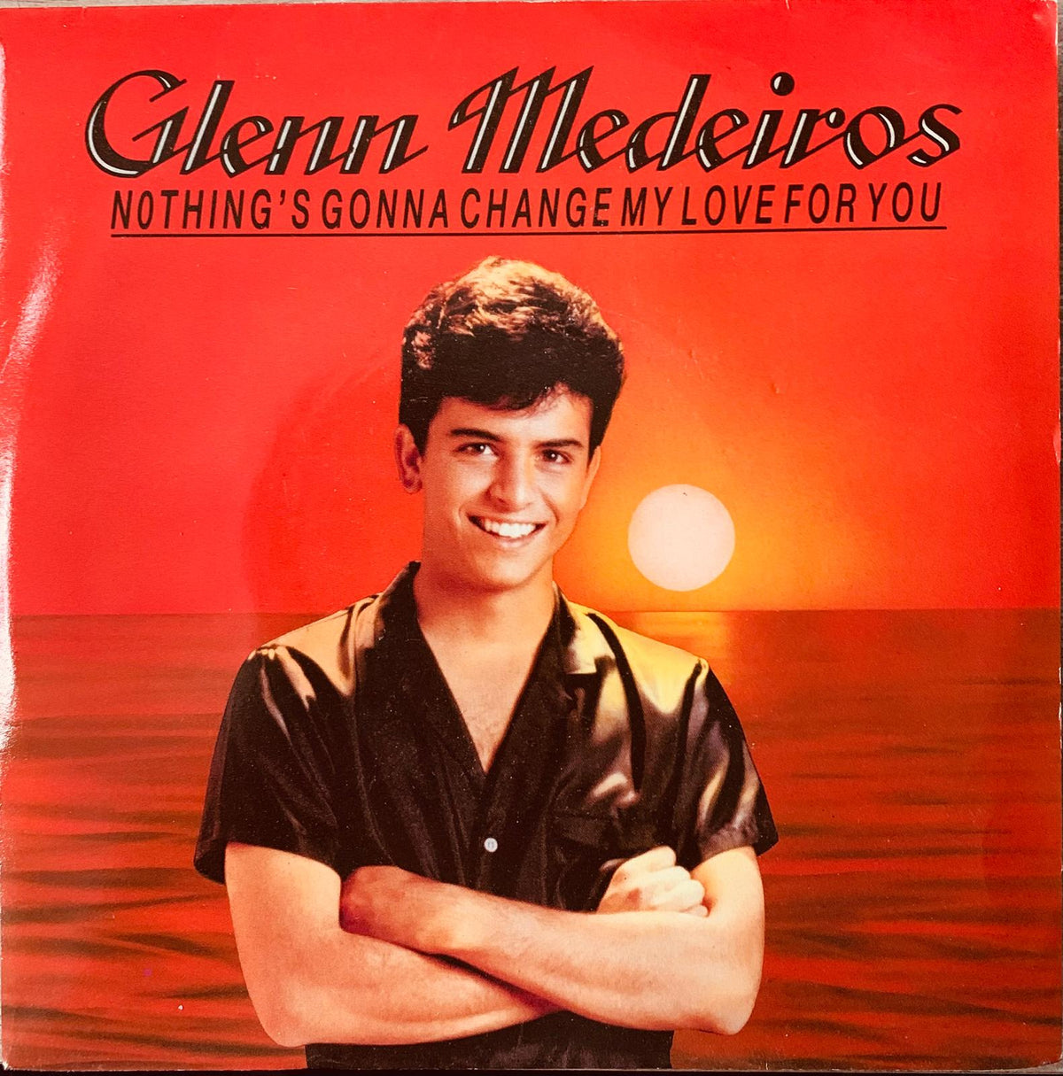 Glenn medeiros wearing black satin shirt with arms crossed against red sunset on original vinyl record cover for nothing’s gonna change my love for you 1987