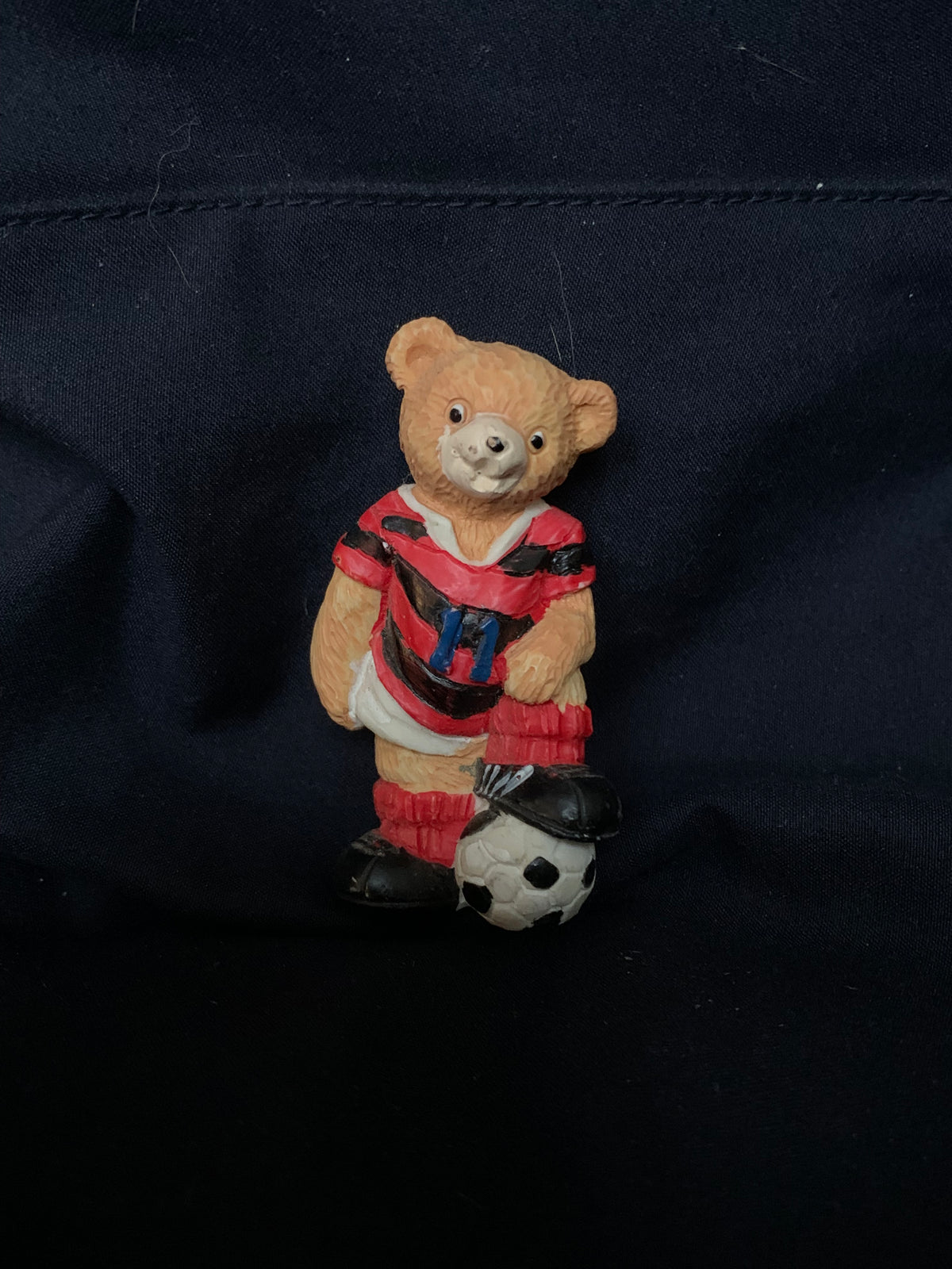 A tan plastic teddy bear figurine in a red and black striped soccer jersey with matching shorts and socks standing next to a black-and-white soccer ball