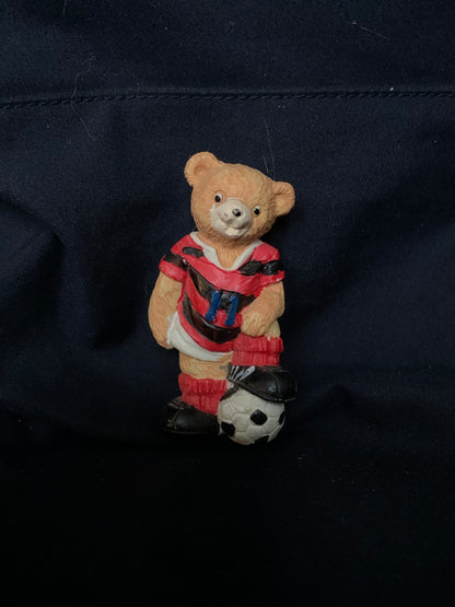A tan plastic teddy bear figurine in a red and black striped soccer jersey with matching shorts and socks standing next to a black-and-white soccer ball
