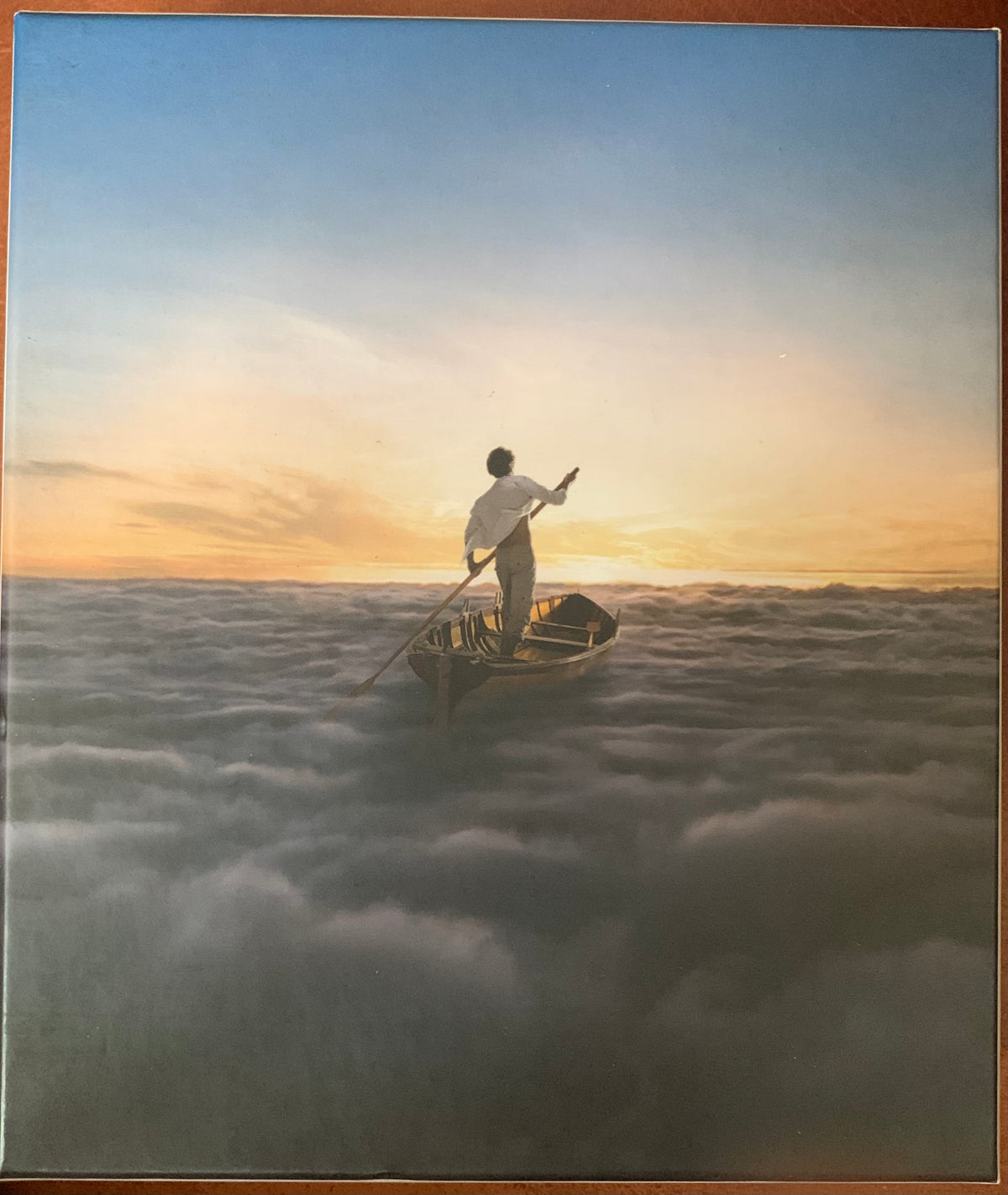 A man in a white shirt stands in a wooden rowboat navigating through clouds at sunset for endless river
