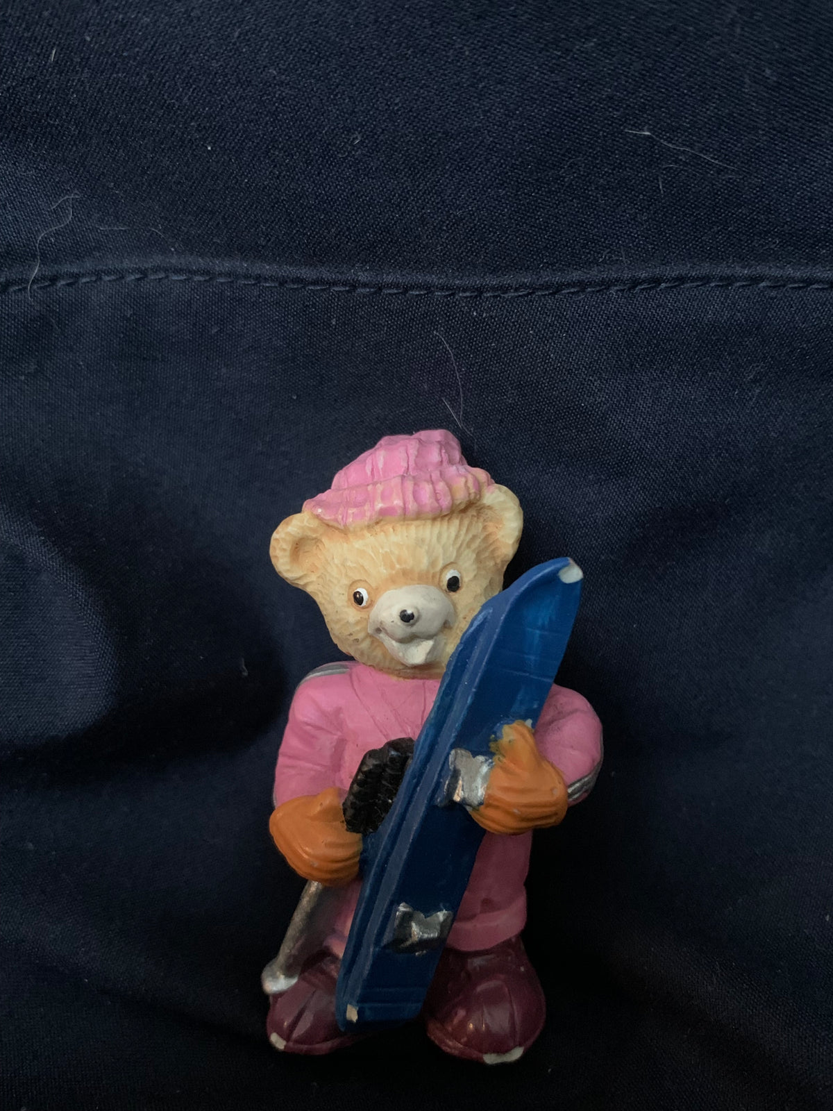 A beige teddy bear figurine wearing a pink knit hat and outfit with orange gloves holding a blue snowboard in toys 001
