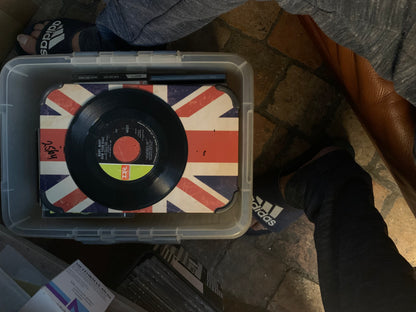 Black vinyl record soul 7 with red center label and green stripe resting on union jack surface in clear storage bin