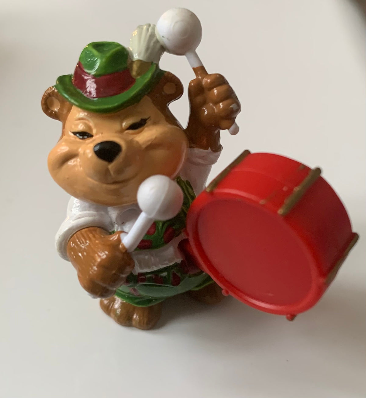 Brown bear figurine wearing green hat with red band and white shirt holding drumsticks next to small red drum