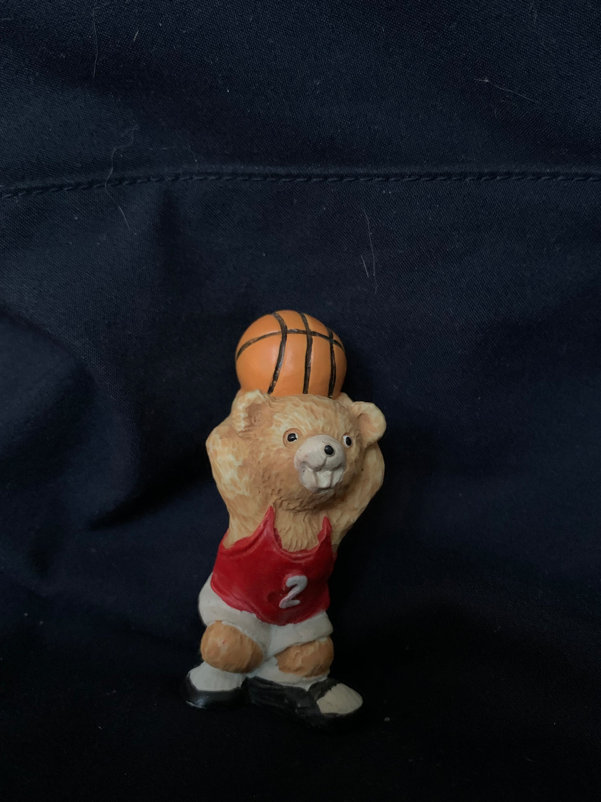 A tan bear figurine toy wearing a red jersey number 2 and white shorts holding an orange basketball on its head