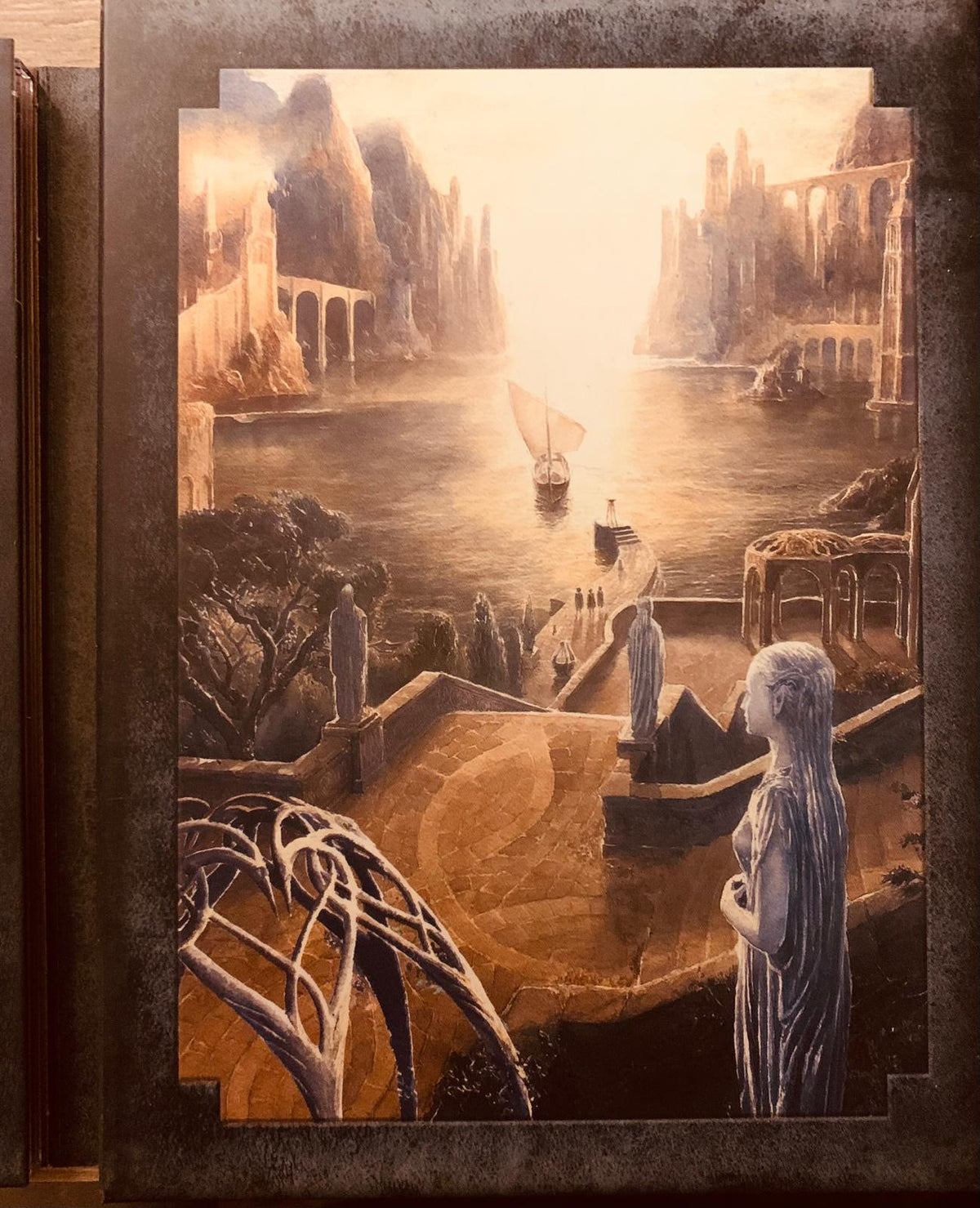 A framed fantasy landscape painting of a golden cityscape with stone arches sailboat and statues in the lord of the rings dvd set