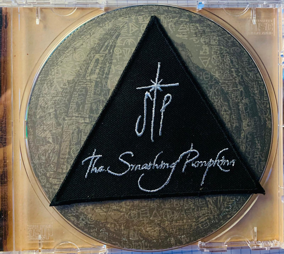 Black triangular patch with white embroidered the smashing pumpkins logo on a circular original cd background