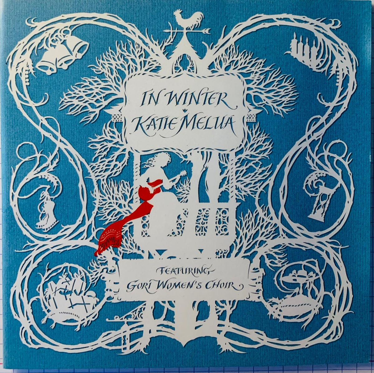 Blue cd album cover of katie melua in winter with intricate white paper-cut art and central red and white figure