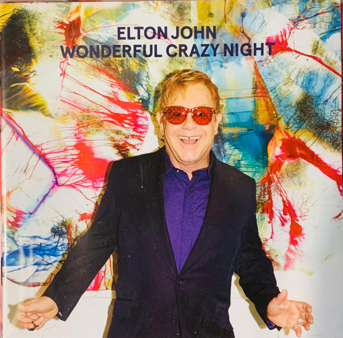 Elton John wearing red tinted sunglasses and a navy suit over a purple shirt on the Wonderful Crazy Night 2016 CD cover
